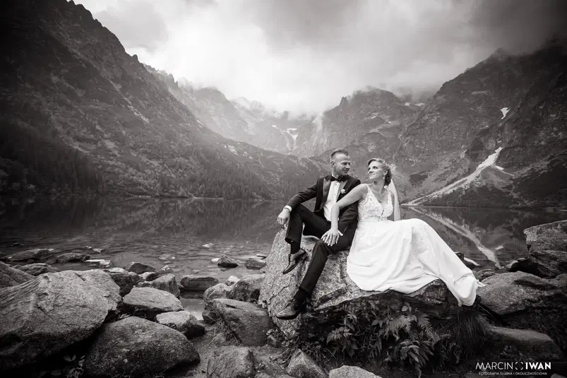  Czarno-białe zdjęcie pary młodej siedzącej na skałach nad jeziorem Morskie Oko w Tatrach, otoczonej majestatycznymi górami i chmurami. 