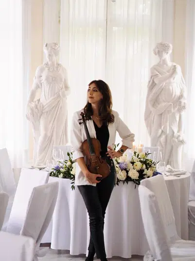 Kobieta z skrzypcami na tle eleganckiej sali z białymi rzeźbami i dekoracjami kwiatowymi. 