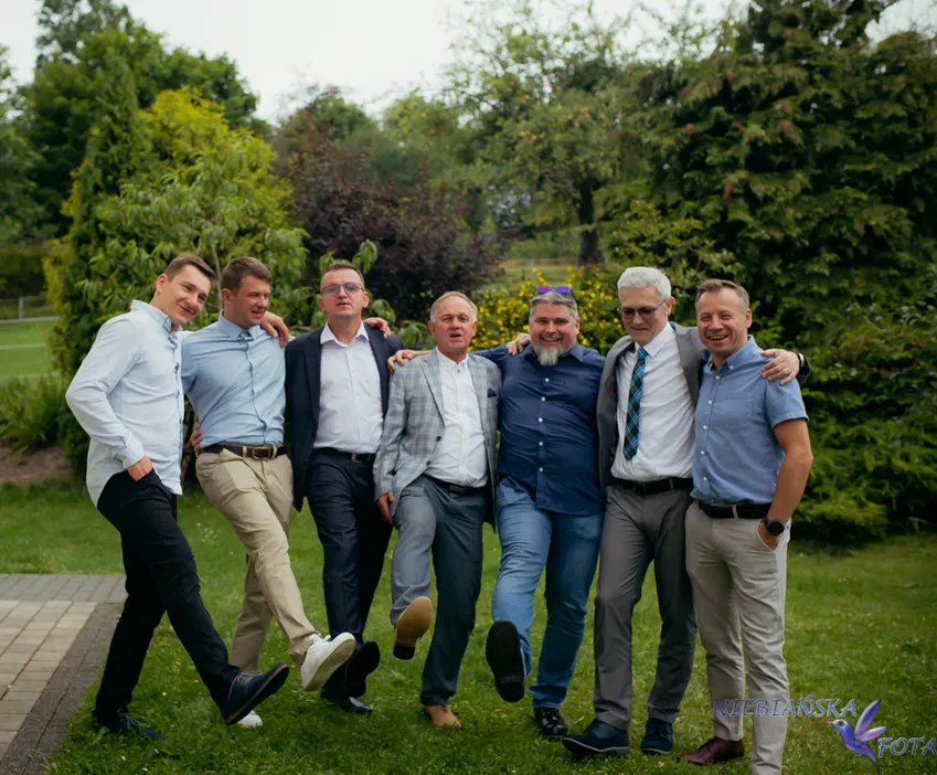 Grupa mężczyzn w eleganckich ubraniach, pozujących razem na świeżym powietrzu w zielonym ogrodzie, na przyjęciu lub weselu.