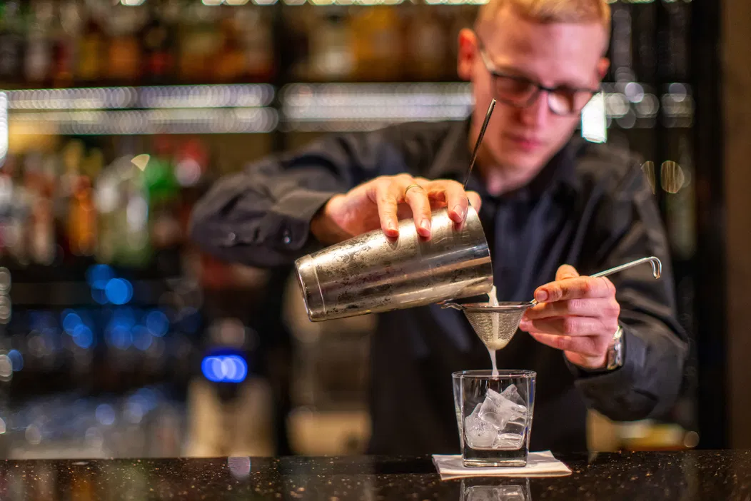 Barman przygotowujący koktajl w eleganckim barze, nalewający płyn przez sitko do szklanki z lodem.