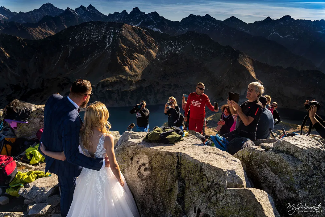 Para młoda na szczycie górskim z fotografami w tle, sceneria Tatr, sesja ślubna na wysokościach.