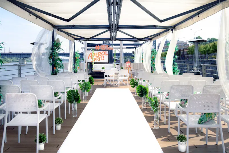  Zadaszona przestrzeń eventowa nad rzeką z białymi krzesłami i roślinami, tło z logiem Aperol Spritz. 