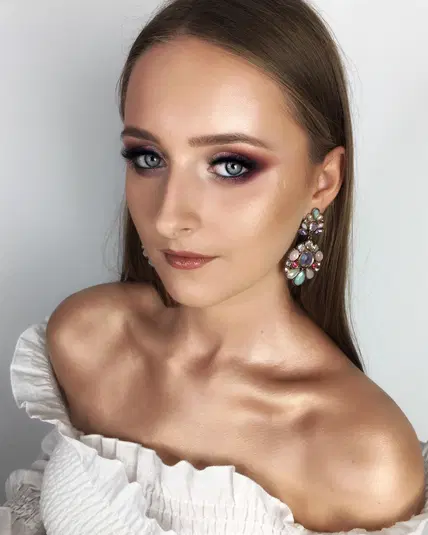  Portret kobiety z eleganckim makijażem, dużymi kolczykami i białą bluzką z odkrytymi ramionami. 