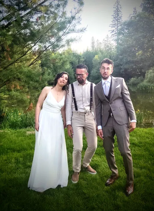 Trzy osoby w eleganckich strojach pozujące na tle zieleni i drzewa w plenerze.