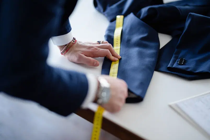  Osoba mierząca rękaw marynarki żółtą miarką krawiecką na tle stołu, elegancki garnitur. 