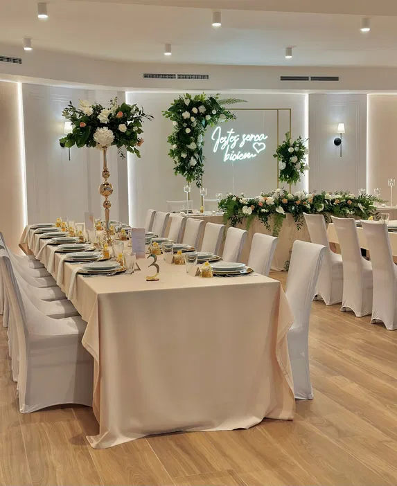 Elegancka sala weselna z elegancko nakrytymi stołami, ozdobiona kwiatami i napisem Jesteś serca biciem na tle ściany. Idealna dekoracja na wesele.