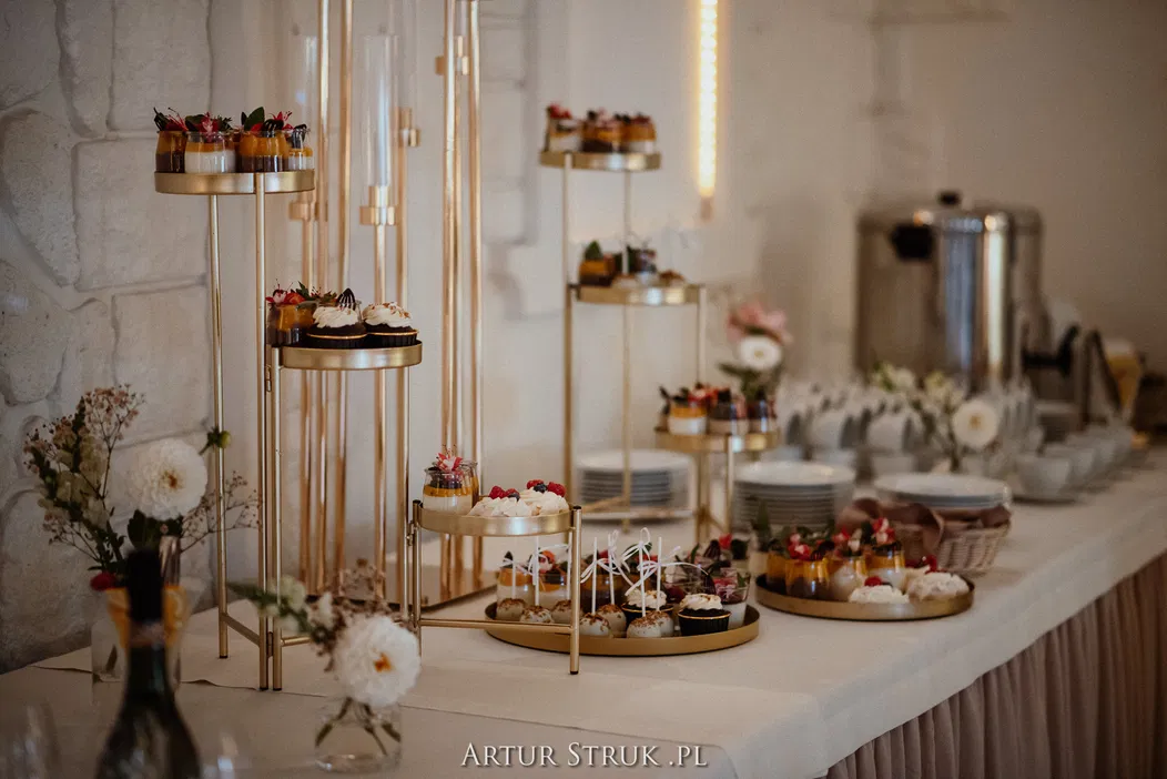 Luksusowy bufet z deserami na złotych stojakach, ozdobiony kwiatami, elegancka dekoracja, catering weselny, ciasta i babeczki na przyjęcie.
