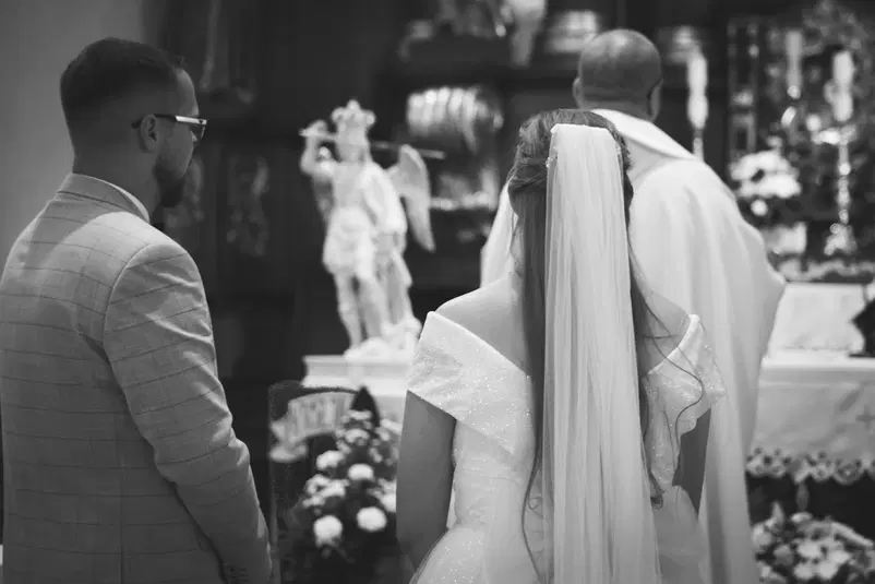  Czarno-białe zdjęcie z ceremonii ślubnej przedstawiające pannę młodą w białej sukni i welonie, stojącą obok pana młodego w garniturze, przed ołtarzem. W tle widoczna figura anioła oraz kwiaty. 