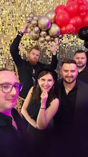  Grupa osób uśmiechających się na tle złotej ściany z balonami w kształcie chmurki w kolorach czerwonym i złotym, ubranych elegancko, impreza lub wydarzenie towarzyskie. 