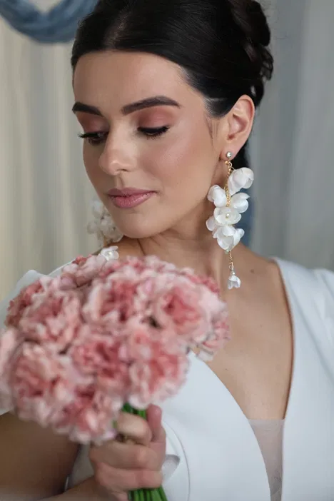 Woman in elegant white dress holding pink flowers, wearing floral earrings, looking down.