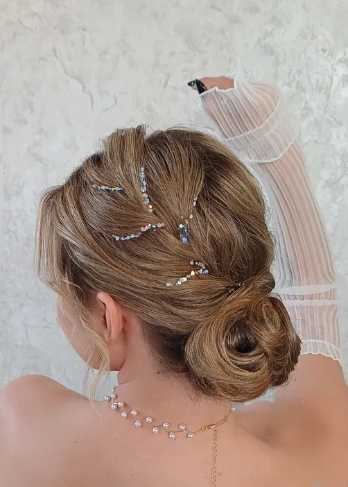 Elegancka fryzura z kokiem i ozdobami z kryształków na włosach blond, stylizacja na uroczystość, biżuteria na szyi.