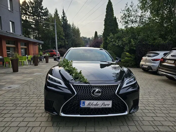  Czarny samochód Lexus ozdobiony zielonym stroikiem, zaparkowany na brukowanym podjeździe przed budynkiem, otoczony przez inne zaparkowane auta i roślinność. 