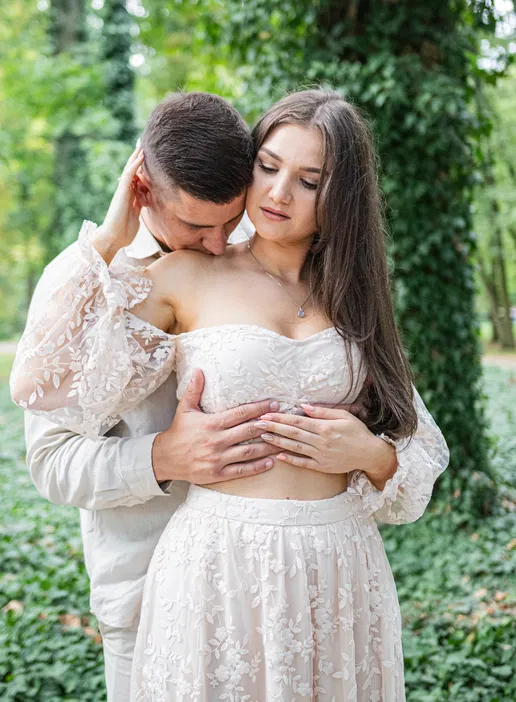 Para w romantycznej pozie w parku, ubrana w eleganckie stroje, otoczona zielenią.