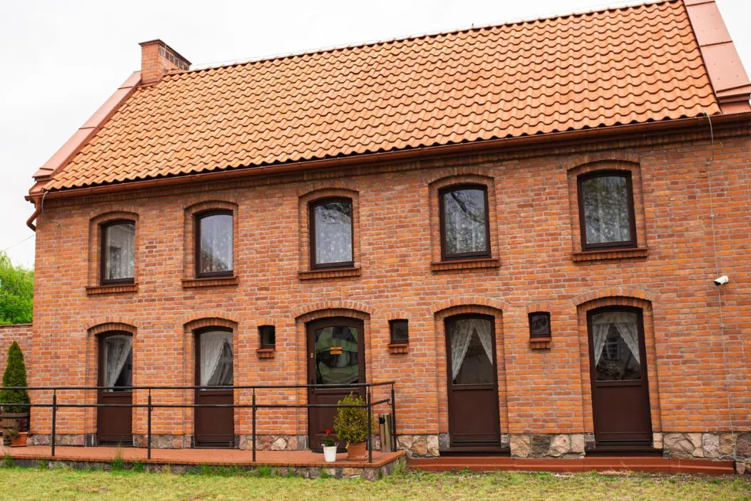 Ceglany dom z dwuspadowym dachem, brązowe drzwi i okna, klasyczna architektura, zielony trawnik, styl wiejski.