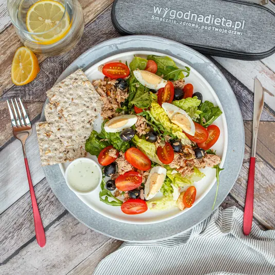  Salata z tuńczykiem, jajkami, pomidorkami koktajlowymi, oliwkami, chrupkim pieczywem i sosem na talerzu, obok karafka z wodą i cytryną oraz zestaw sztućców. Idealny posiłek dla diety pudełkowej od wygodnadieta.pl. 