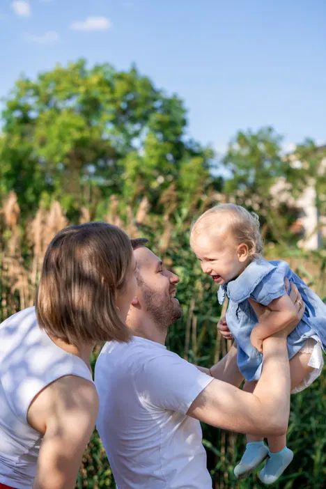Rodzina na spacerze w parku, tata bawiący się z uśmiechniętym dzieckiem na tle zieleni i błękitnego nieba.