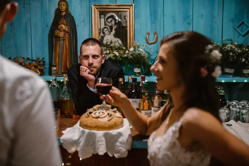  Mężczyzna w garniturze przy barze z alkoholem, skupiony na parze weselnej z tradycyjnym chlebem weselnym w tle, ozdobnym wystrojem i obrazem religijnym na niebieskim drewnianym tle. 
