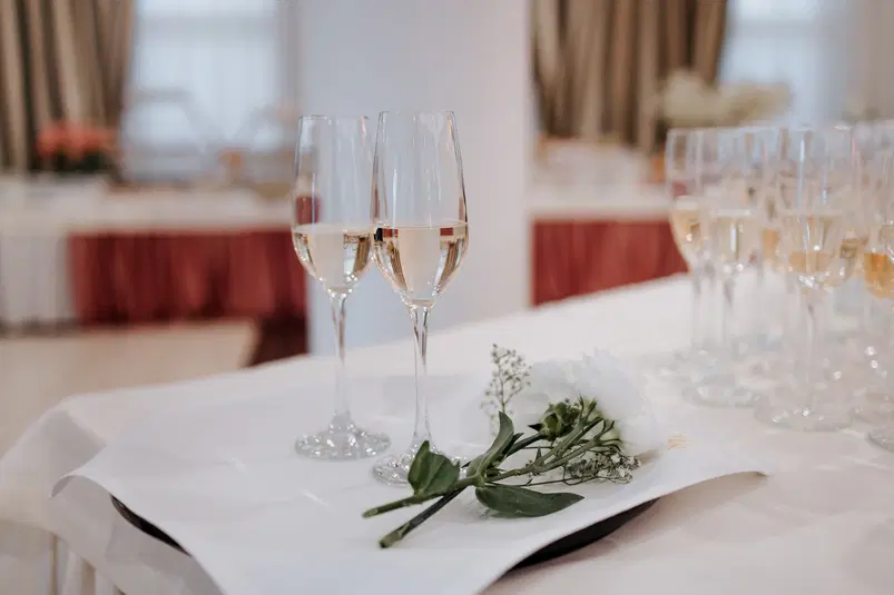  Dwa kieliszki szampana i biała róża na elegancko nakrytym stole - wesele, przyjęcie, celebracja. 