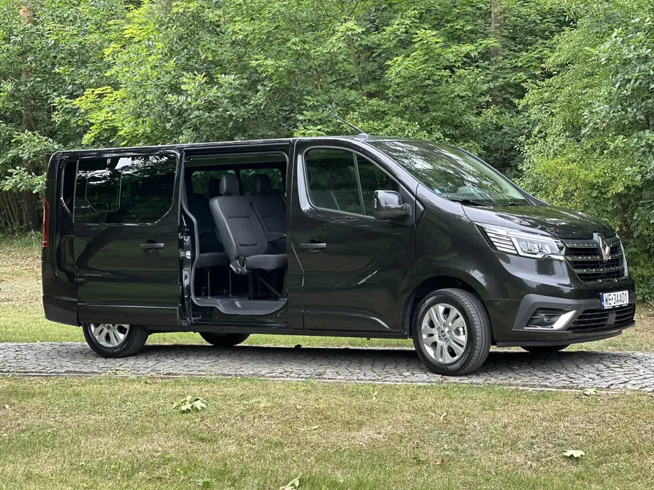 Czarny minivan typu Renault Trafic z otwartymi drzwiami bocznymi, stojący na tle zielonego lasu.