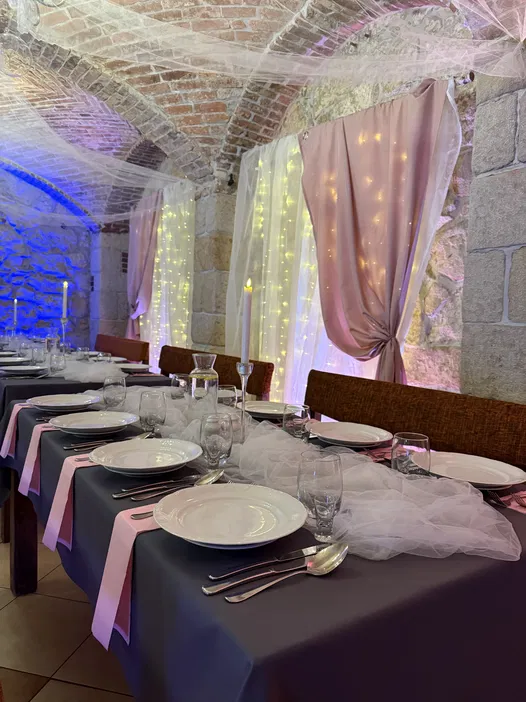 Sala weselna z ceglaną sklepką, udekorowana stołami z szarymi obrusami, różowymi serwetkami i białymi talerzami, w tle podświetlone zasłony i kamienne ściany.