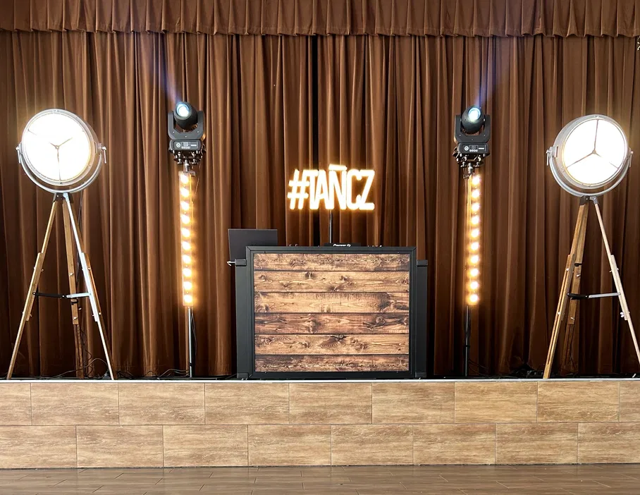 DJ stanowisko z drewnianym blatem, oświetlone reflektorami, z neonowym napisem TAŃCZ na tle brązowej zasłony. Idealne miejsce na event muzyczny.
