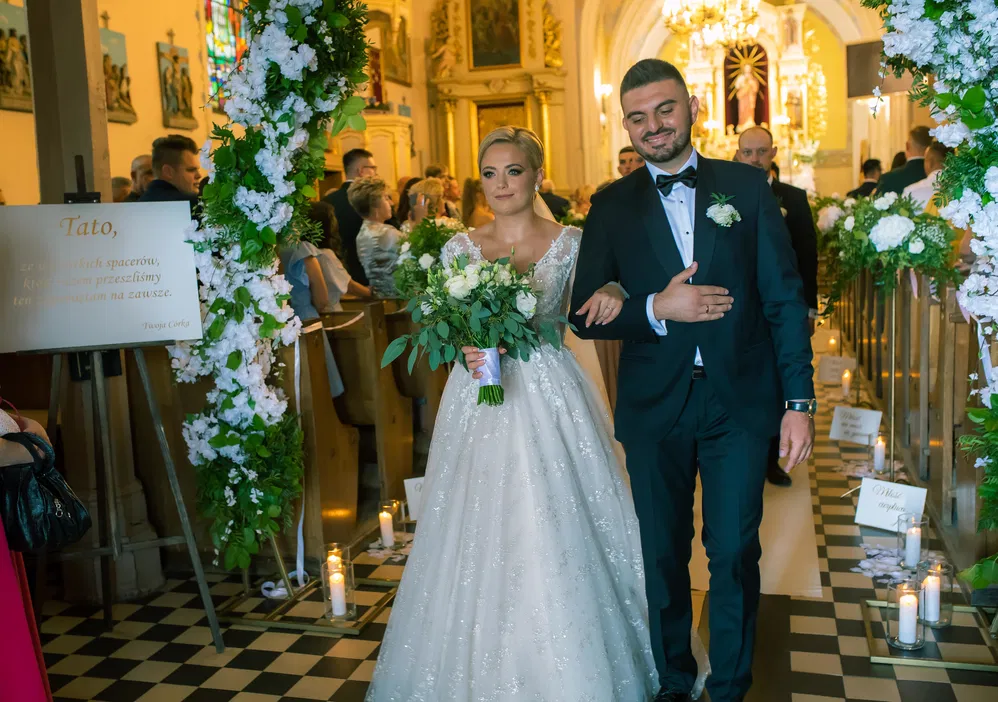 Para młoda wychodzi z kościoła po ceremonii ślubnej, ozdobiona kwiatami i świecami na tle gości.