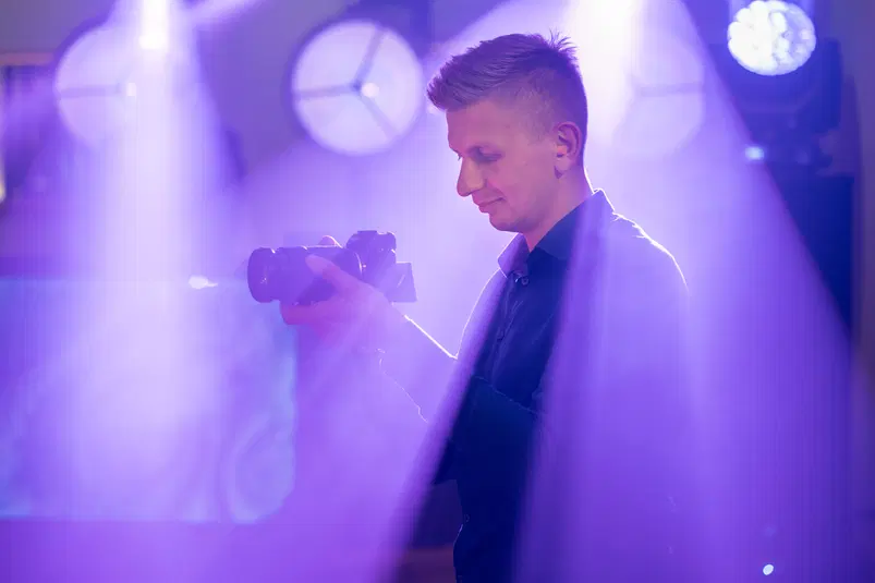  Osoba trzymająca aparat fotograficzny na imprezie z fioletowym oświetleniem w tle. 