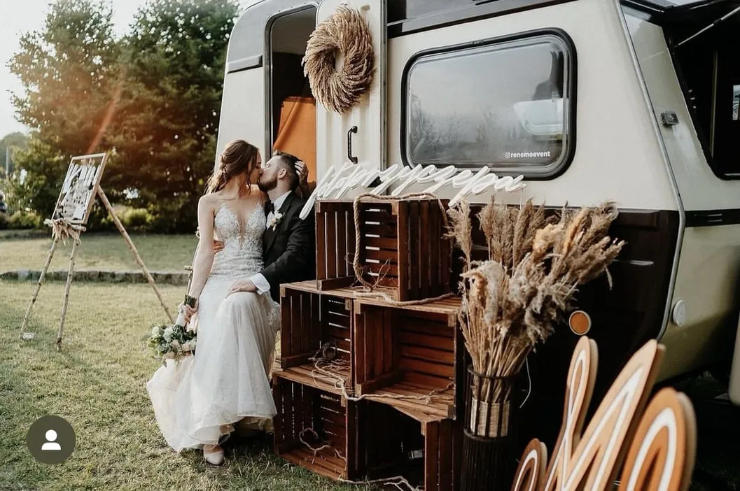 Para młoda całująca się przed rustykalną przyczepą na świeżym powietrzu, otoczona dekoracjami ślubnymi w stylu boho, z drewnianymi skrzynkami i suszonymi kwiatami.
