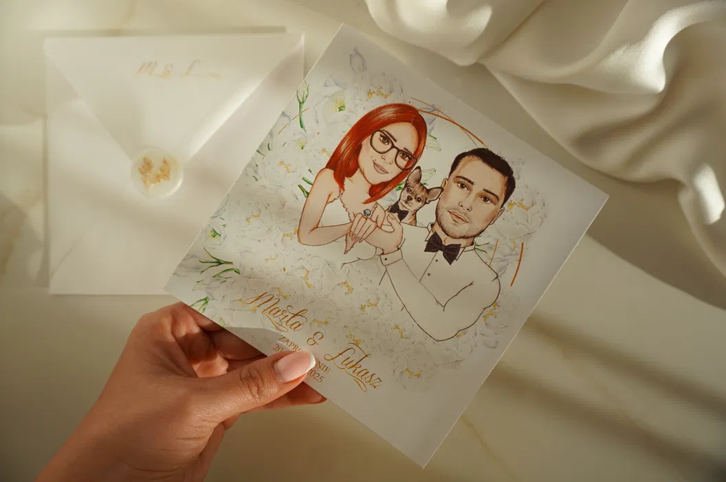 Ręka trzymająca zaproszenie ślubne z ilustracją pary i psem, koperta w tle, eleganckie detale.