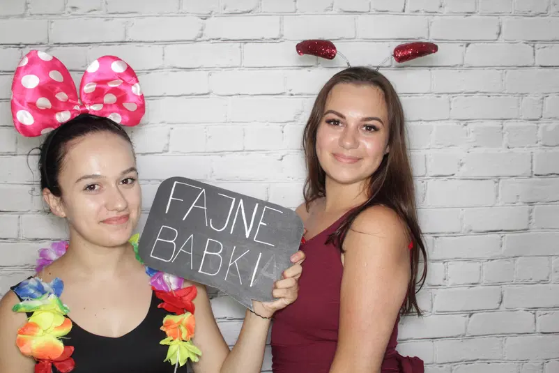  Dwie kobiety z akcesoriami do fotobudki, jedna trzyma tabliczkę z napisem Fajne Babki, tło z białych cegieł. 