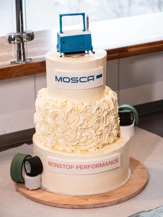 Dwupiętrowy tort dekorowany kwiatami z logo MOSCA i napisem NONSTOP PERFORMANCE, na szczycie tortu niebieska dekoracja w kształcie maszyny, leżące obok rolki w różnych kolorach, stojący na drewnianej desce.