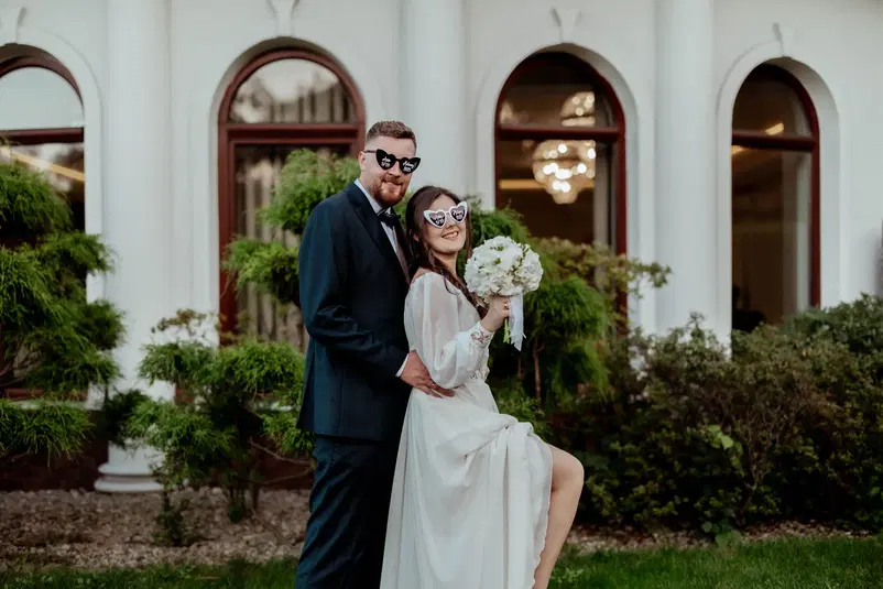  Para młoda w okularach przeciwsłonecznych, pozująca przed eleganckim budynkiem; panna młoda w białej sukni trzyma bukiet kwiatów. 