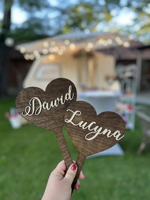 Drewniane serca z imionami Dawid i Lucyna, dekoracja weselna na tle rozmazanych światełek ogrodowych.