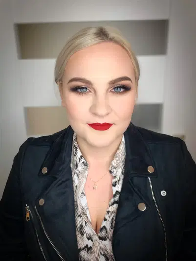  Blondynka o niebieskich oczach, z wyrazistym makijażem oczu i czerwonymi ustami, ubrana w czarną kurtkę i wzorzystą bluzkę, patrzy prosto w obiektyw. 