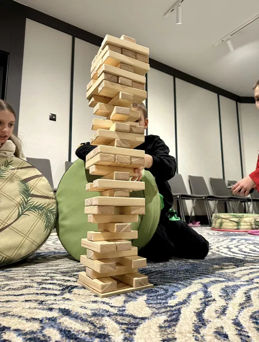 Gra Jenga w trakcie budowy, dzieci koncentrują się na stabilnej wieży z drewnianych klocków, tło z wzorzystym dywanem i nowoczesnymi krzesłami.