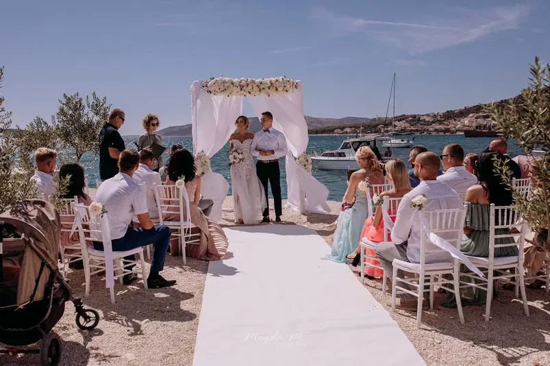  Ceremonia ślubna na plaży, para młoda pod kwiatowym łukiem, goście siedzący na białych krzesłach, morze i żaglówki w tle. 