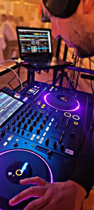 DJ przy konsoli mikserskiej w klubie nocnym, na pierwszym planie gramofon z kolorowym podświetleniem i laptop z oprogramowaniem do miksowania muzyki w tle.
