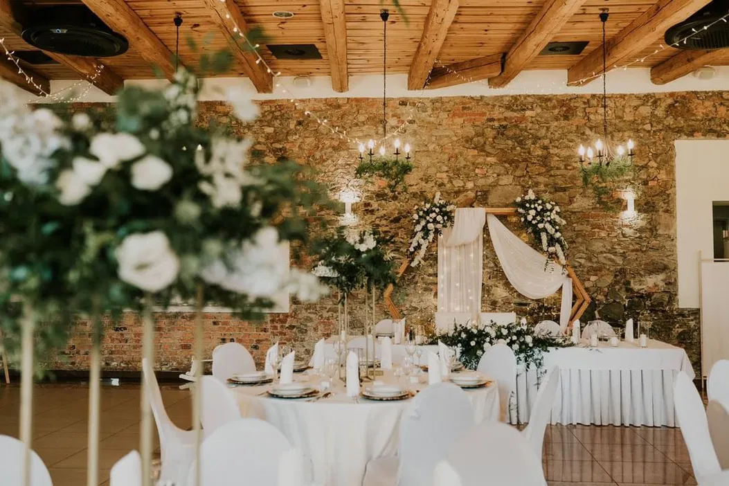 Elegancka sala weselna z rustykalnym wystrojem, drewnianymi belkami na suficie i kamiennymi ścianami, ozdobiona białymi kwiatami i świecznikami, przygotowana na przyjęcie weselne.
