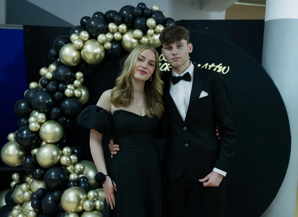 Para młodych ludzi ubrana elegancko, kobieta w czarnej sukni, mężczyzna w czarnym garniturze i muszce, stojących przy dekoracji z czarnych i złotych balonów.