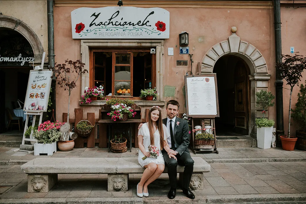 Para młoda siedząca przed restauracją Zachcianek w klimatycznym, zabytkowym otoczeniu, ozdobioną kolorowymi kwiatami.