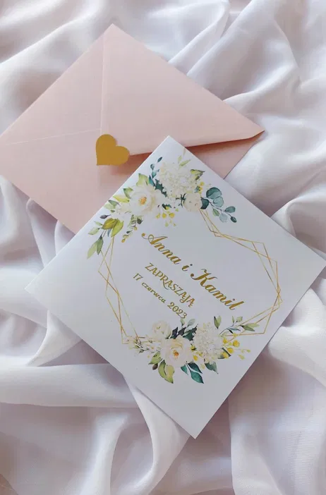 Zaproszenie ślubne z eleganckim kwiatowym wzorem i personalizowanym tekstem na tle jasnej tkaniny, obok pastelowej koperty z sercem.
