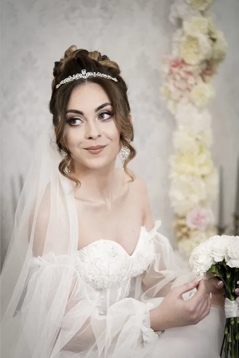 Młoda panna młoda w eleganckiej sukni ślubnej z diademem i bukietem kwiatów na tle dekoracji z jasnych kwiatów.