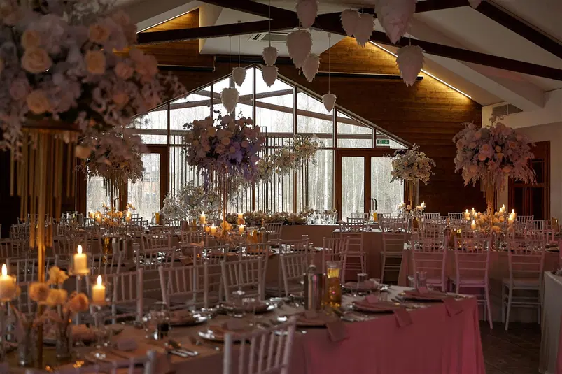  Elegancka sala weselna z dekoracjami kwiatowymi i świecami, przygotowana na przyjęcie, z widokiem na duże okna i drewniane wykończenia. 