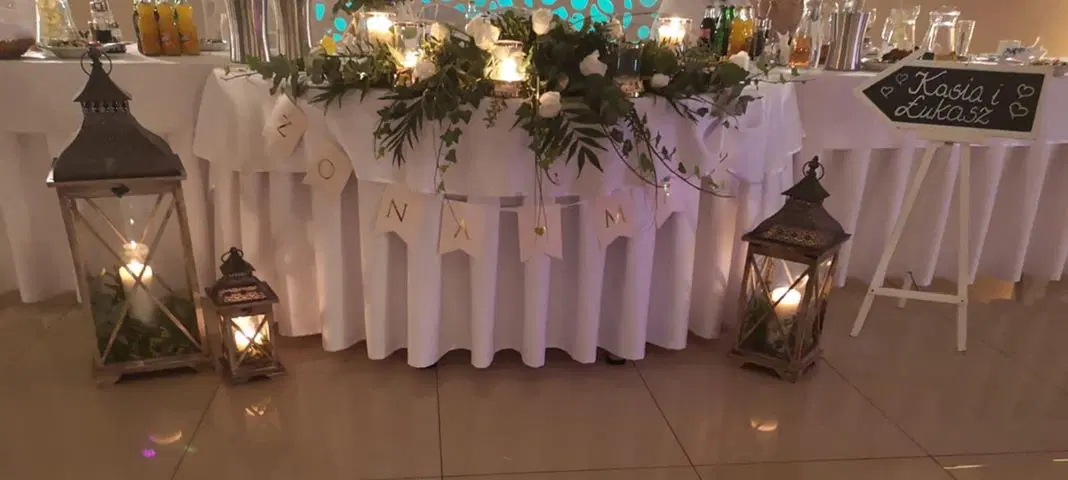  Stylowe dekoracje stołu weselnego z zielenią i świecami, urocze latarenki oraz tablica z imionami Kasia i Łukasz, elegancka atmosfera przyjęcia. 