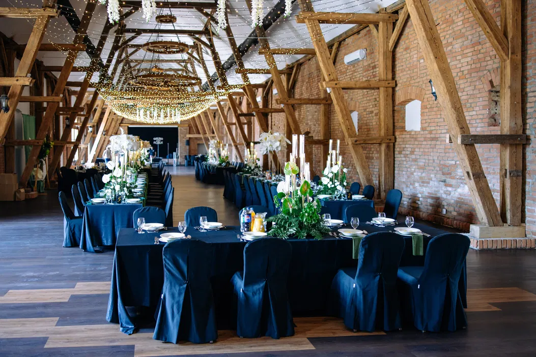 Elegancka sala bankietowa z drewnianą konstrukcją, ciemnymi krzesłami i stołami ozdobionymi zielenią i świecami, przygotowana na przyjęcie weselne.