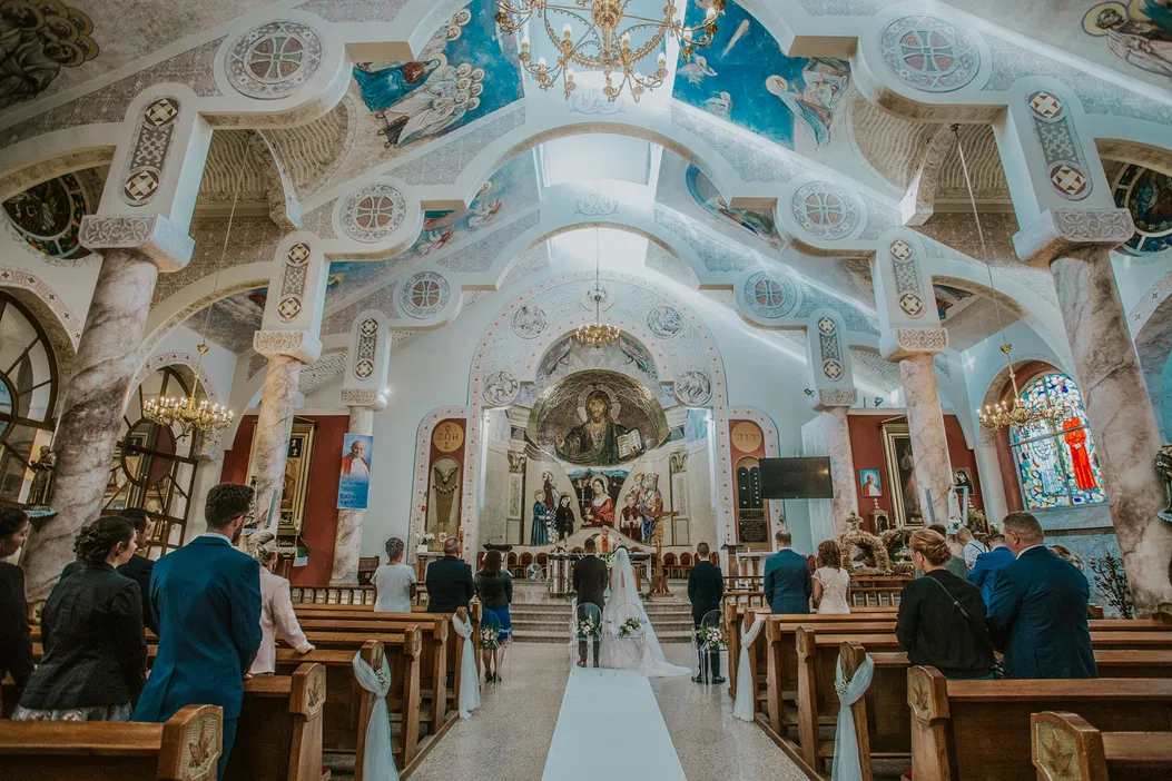 Wnętrze kościoła podczas ceremonii ślubnej, piękne dekoracje i freski, młoda para stojąca przy ołtarzu, goście zgromadzeni w ławkach.