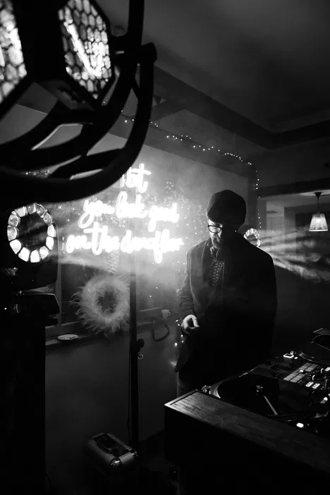 Czarnobiała fotografia DJ-a z neonowym napisem, w klubowej atmosferze z efektami dymu i światła.