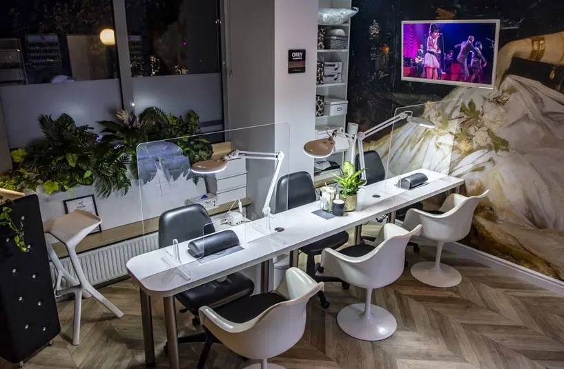  Nowoczesne wnętrze salonu kosmetycznego z eleganckimi stanowiskami do manicure, w tle telewizor z występem scenicznym, bogata roślinność i luksusowe wykończenia. 