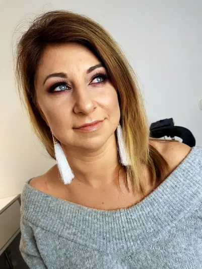  Nieznana kobieta w szarym swetrze z dużymi białymi kolczykami i makijażem oczu, patrząca w bok, w jasnym pomieszczeniu. 