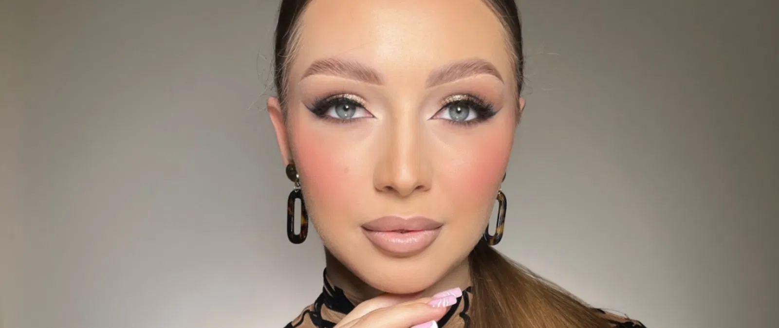 Nieznana kobieta z makijażem glamour, z brązowymi włosami związanymi w kucyk, z dużymi kolczykami, pozująca do zdjęcia.